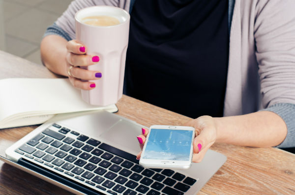 Ung kvinnan med en mugg kaffe i ena handen. I andra handen en mobiltelefon. Hon sitter framför en dator. 
